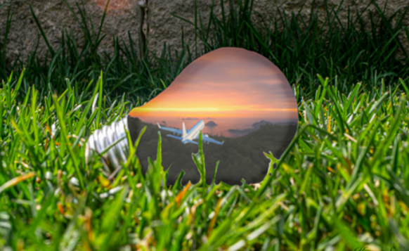 Colour photo of a lightbulb on grass. Inside the lightbulb is a sunset sky and an airplane