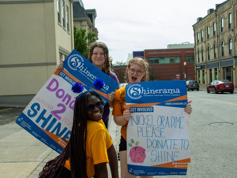 two Laurier students each hold a poster for Shinerama. The poster on the left says Donate 2 Shine and the one on the right says nickel or a dime donate to shrine. A student in a yellow shirt crouches down in fron of the signs